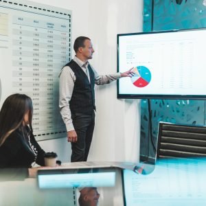 A business meeting in a boardroom: a caucasian man entrepreneur is pointing on a plasma screen on the wall with a chart of profit indicators while his female colleague is sitting at the table aloof