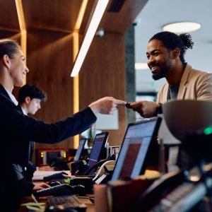 Happy receptionist giving room cardkey to African American hotel guest at reception desk.